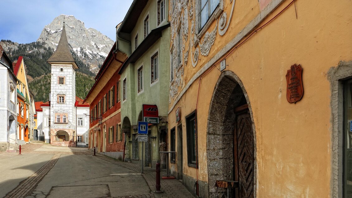 Ein historisches Gebäude mit dekorativen Fassadenmalereien und Fenstern. Im Hintergrund sind Berge zu sehen, bei klarem Himmel. | © Pezmann