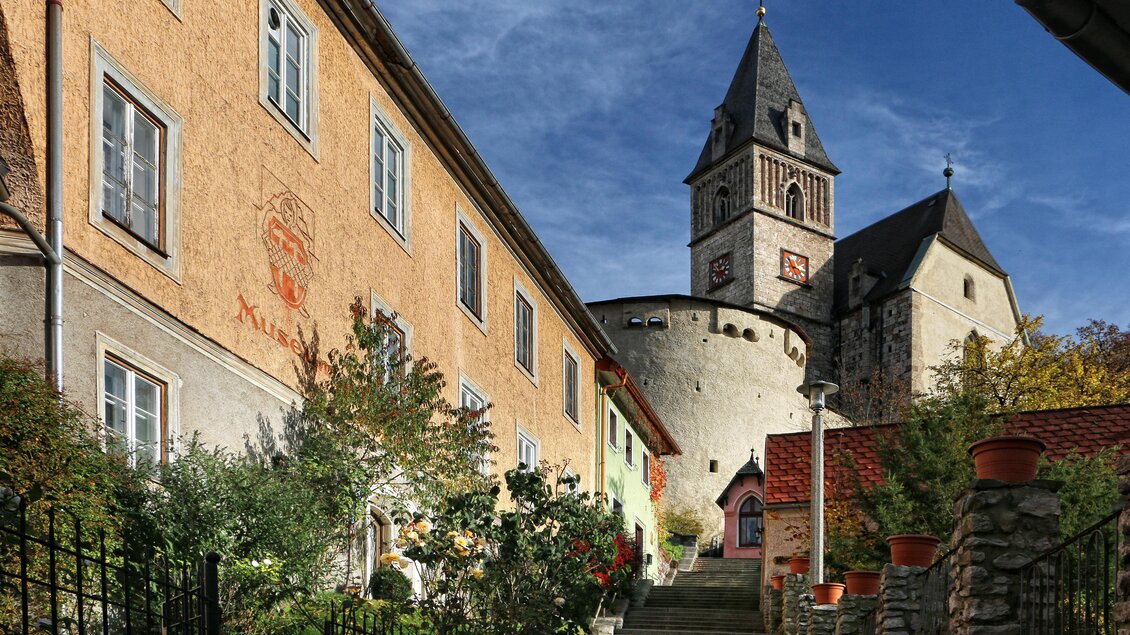 Ein malerischer Dorfweg mit alten Häusern und einer Kirche im Hintergrund. Der Himmel ist blau und die Landschaft ist grün und einladend. | © Pezmann