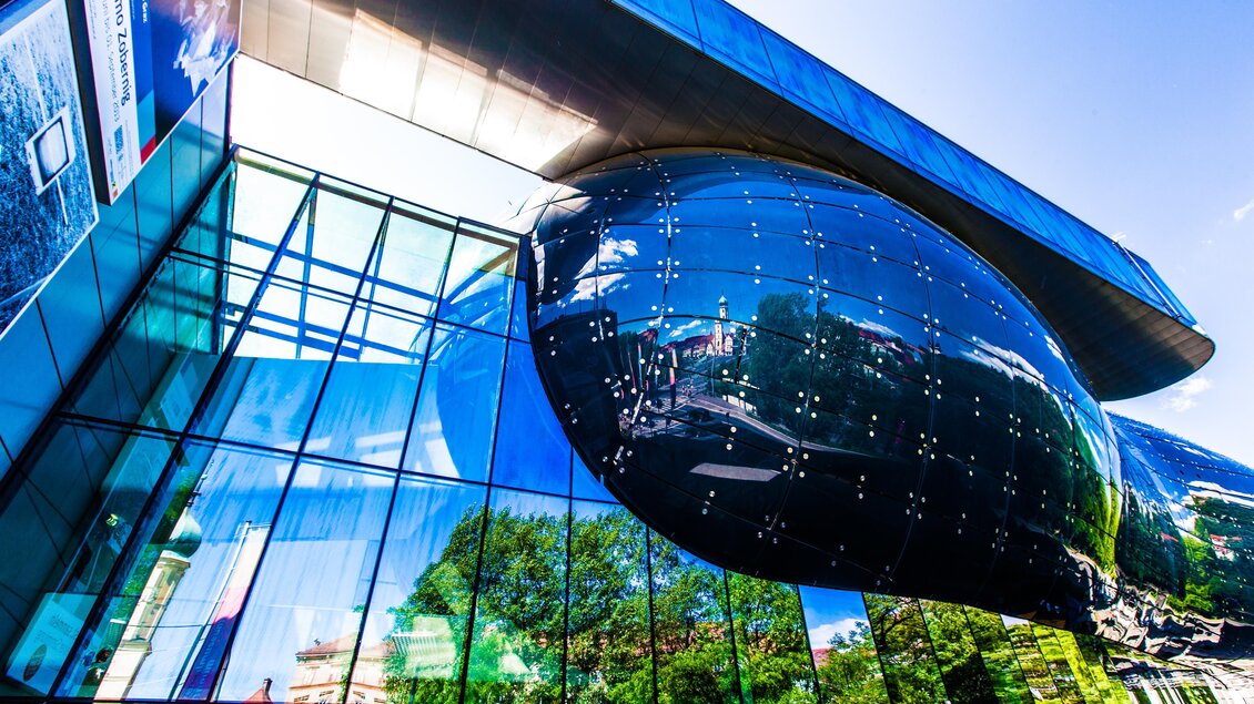 Ein modernes Gebäude mit einer glänzenden, ovoiden Form und großen Glasflächen. Die reflektierende Fassade spiegelt die Umgebung wider. | © Graz Tourismus - Harry Schiffer