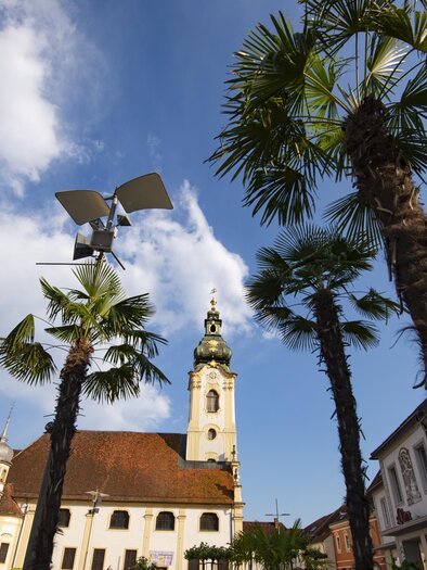 Ein schöner Platz mit Palmen und historischen Gebäuden. Im Hintergrund ist ein Kirchturm unter einem blauen Himmel zu sehen. | © Oststeiermark Tourismus
