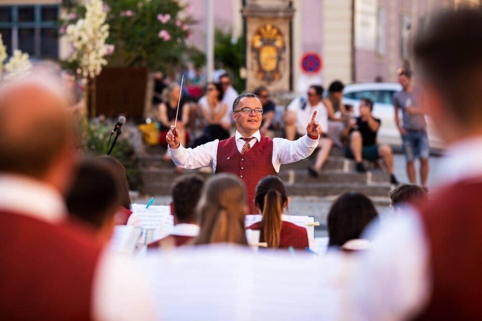 Ein Dirigent leitet ein Orchester bei einem Auftritt im Freien. Im Hintergrund sind Zuschauer zu sehen, die der Musik lauschen.