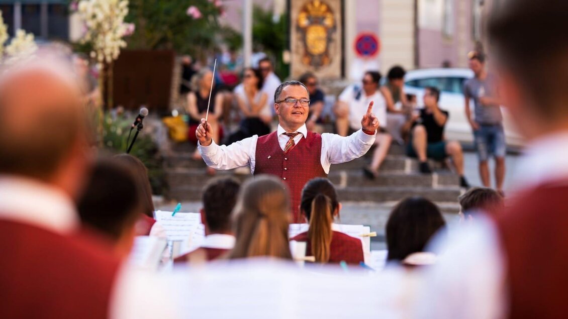 Ein Dirigent leitet ein Orchester bei einem Auftritt im Freien. Im Hintergrund sind Zuschauer zu sehen, die der Musik lauschen.