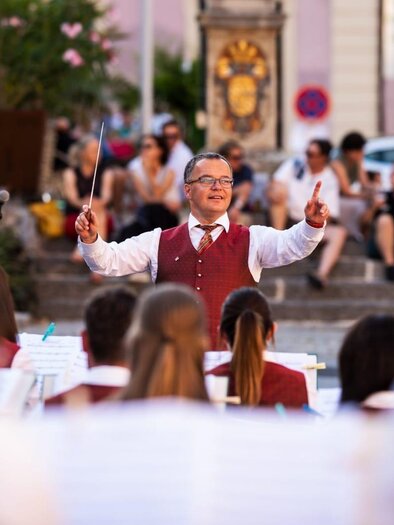 Ein Dirigent leitet ein Orchester bei einem Auftritt im Freien. Im Hintergrund sind Zuschauer zu sehen, die der Musik lauschen.