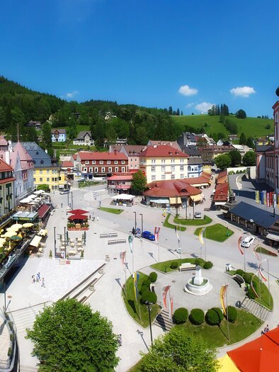 Ein malerischer Marktplatz mit bunten Gebäuden und einer beeindruckenden Kirche im Hintergrund. Die Umgebung ist grün und einladend, ideal für einen Spaziergang. | © Fred Lindmoser