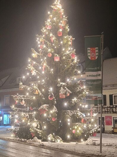 Ein festlich geschmückter Weihnachtsbaum mit Lichtern und Ornamenten steht auf einem schneebedeckten Platz. Es ist Nacht und die Umgebung ist ruhig und winterlich. | © TV Südsteiermark - Irene Löschnig
