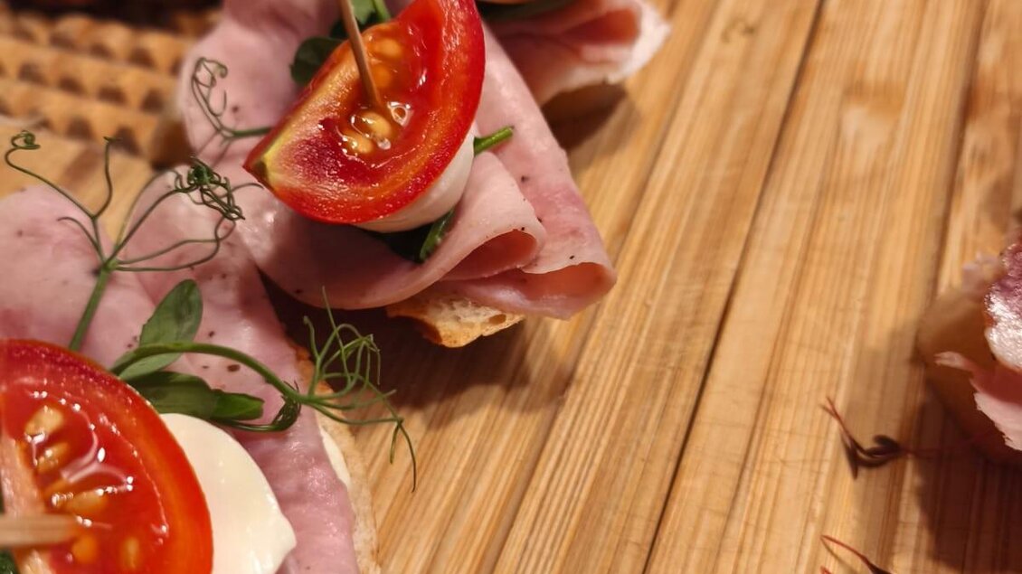 Eine appetitliche Platte mit belegten Brötchen, dekoriert mit Schinken, Tomaten und frischen Kräutern. Die Snacks sind kunstvoll angerichtet auf einem Holzbrett. | © Vladi Slodnjak