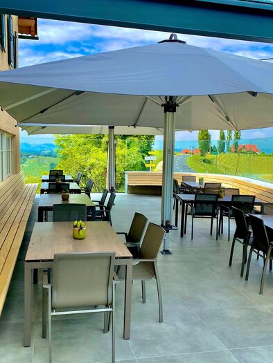 An inviting terrace with tables and chairs under a large sun umbrella. In the background, green meadows and a blue sky can be seen. | © Eckbergerhof