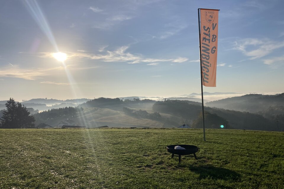 Die aufgehende Sonne taucht die sanften Hügel in warmes Licht und lässt Nebelschwaden über der Landschaft von Edelsbach bei Feldbach schweben. Die Fahne des Steinbüchlers weist den Weg zu einem Naturerlebnis mit herrlichen Ausblicken und regionalem Genuss. | © Verein VBG Steinbüchl