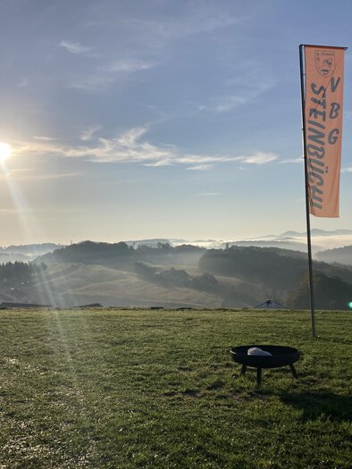 Die aufgehende Sonne taucht die sanften Hügel in warmes Licht und lässt Nebelschwaden über der Landschaft von Edelsbach bei Feldbach schweben. Die Fahne des Steinbüchlers weist den Weg zu einem Naturerlebnis mit herrlichen Ausblicken und regionalem Genuss. | © Verein VBG Steinbüchl