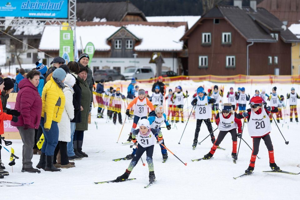 Frontansicht der jungen Langlaufnachwuchstalente beim Kids-Race. Im Hintergrund sind die Häuser von Bad Mitterndorf zu sehen, während entlang der Loipe zahlreiche begeisterte Zuschauer stehen und die jungen Sportler anfeuern. | © Steiralauf/Martin Huber