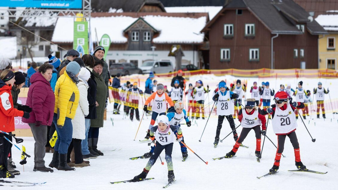Frontansicht der jungen Langlaufnachwuchstalente beim Kids-Race. Im Hintergrund sind die Häuser von Bad Mitterndorf zu sehen, während entlang der Loipe zahlreiche begeisterte Zuschauer stehen und die jungen Sportler anfeuern. | © Steiralauf/Martin Huber