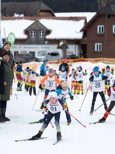 Front view of the young cross-country skiing talents at the kids' race. In the background, the houses of Bad Mitterndorf can be seen, while numerous enthusiastic spectators line the trail and cheer on the young athletes. | © Steiralauf/Martin Huber
