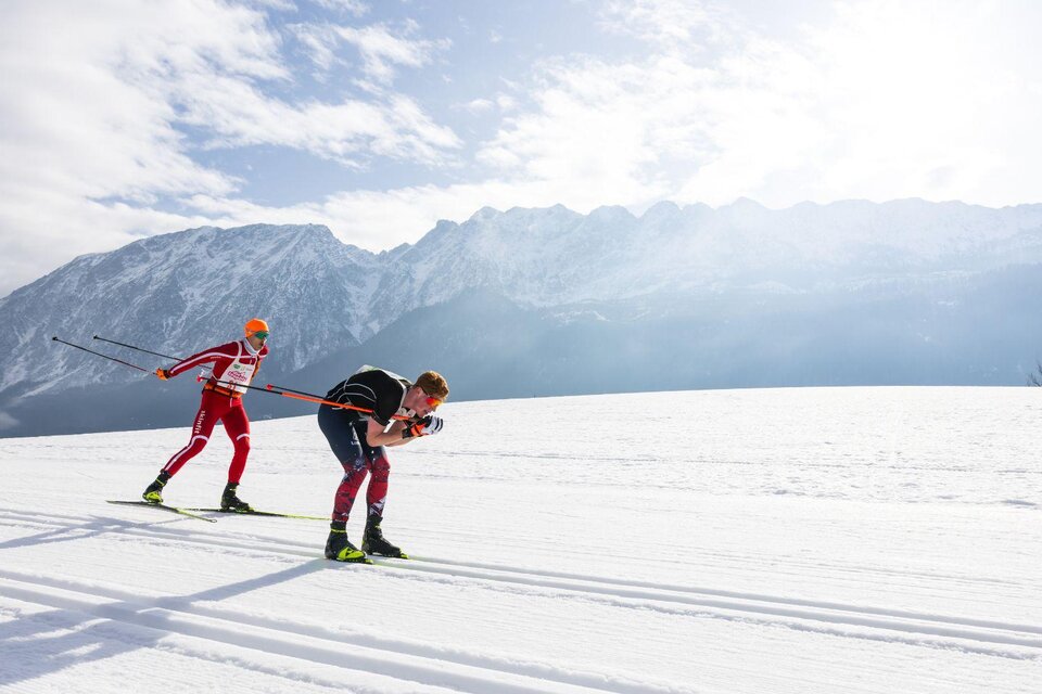 Zwei Langläufer sind auf der Loipe unterwegs, einer in klassischer Technik, der andere beim Skaten. Beide sind fokussiert auf das Rennen, während im Hintergrund der imposante, winterliche Grimming die verschneite Landschaft prägt. | © Steiralauf/Martin Huber