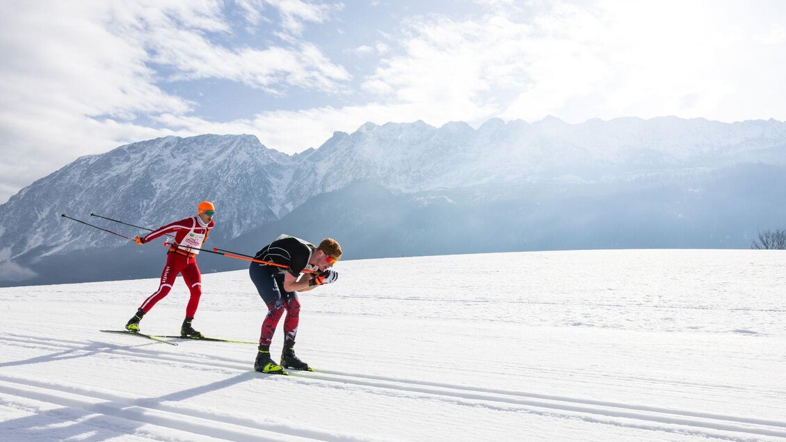 Zwei Langläufer sind auf der Loipe unterwegs, einer in klassischer Technik, der andere beim Skaten. Beide sind fokussiert auf das Rennen, während im Hintergrund der imposante, winterliche Grimming die verschneite Landschaft prägt. | © Steiralauf/Martin Huber