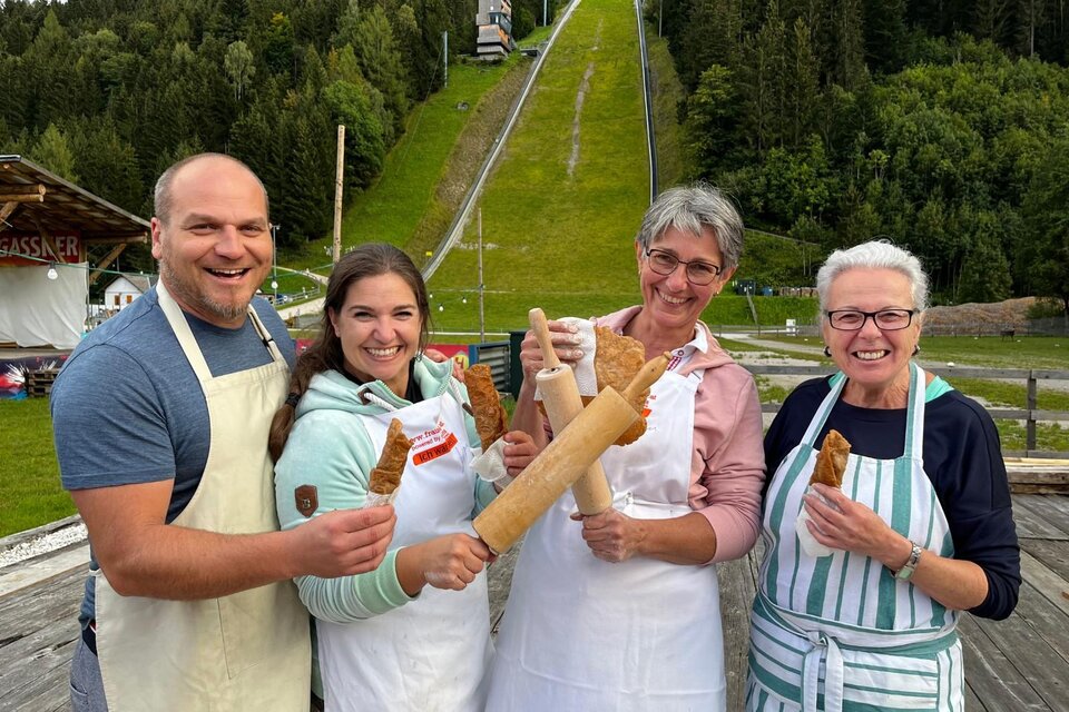 Das Team vom Steirerkrapfenfrühschoppen steht vor der Skiflugschanze Kulm, umgeben von grünen Bäumen, und hält verschiedene Backutensilien. | © Auszeit Ausseerland