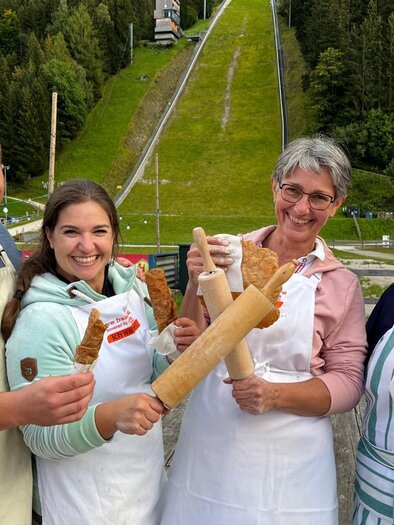 The team from the culinary morning pint "Steirerkrapfenfrühschoppen" stands in front of the Kulm ski flying ramp, surrounded by green trees, and holds various baking utensils. | © Auszeit Ausseerland