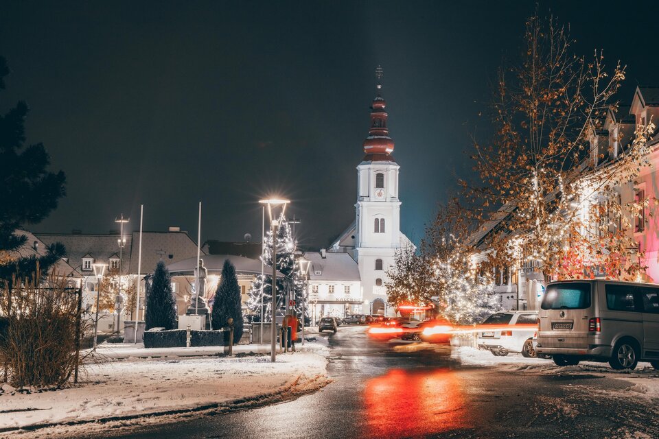 Winterliche Abendstimmung in Fehring mit verschneitem Hauptplatz, festlich beleuchteten Bäumen und der Pfarrkirche Fehring im Hintergrund. | © Sebastian Friedl
