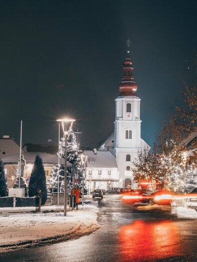 Wintry evening atmosphere in Fehring with a snow-covered main square, festively lit trees, and the parish church of Fehring in the background. | © Sebastian Friedl