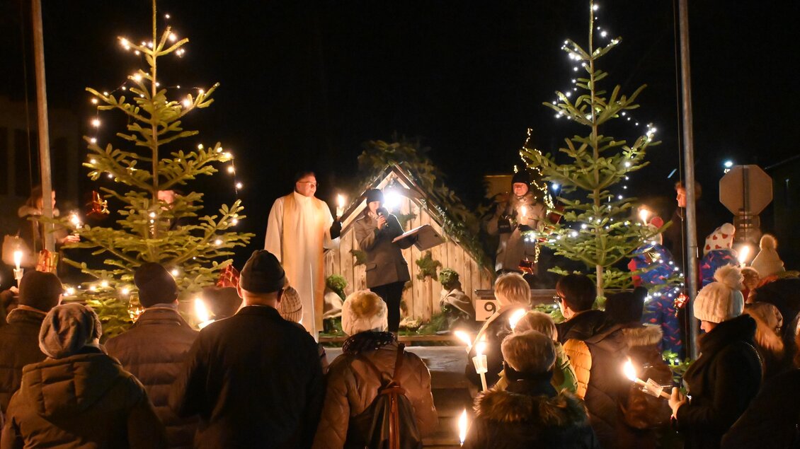 Eine festliche Versammlung bei Dunkelheit mit Menschen, die Kerzen halten. Im Hintergrund stehen geschmückte Tannenbäume und eine Weihnachtskrippe. | © Steirische Roas