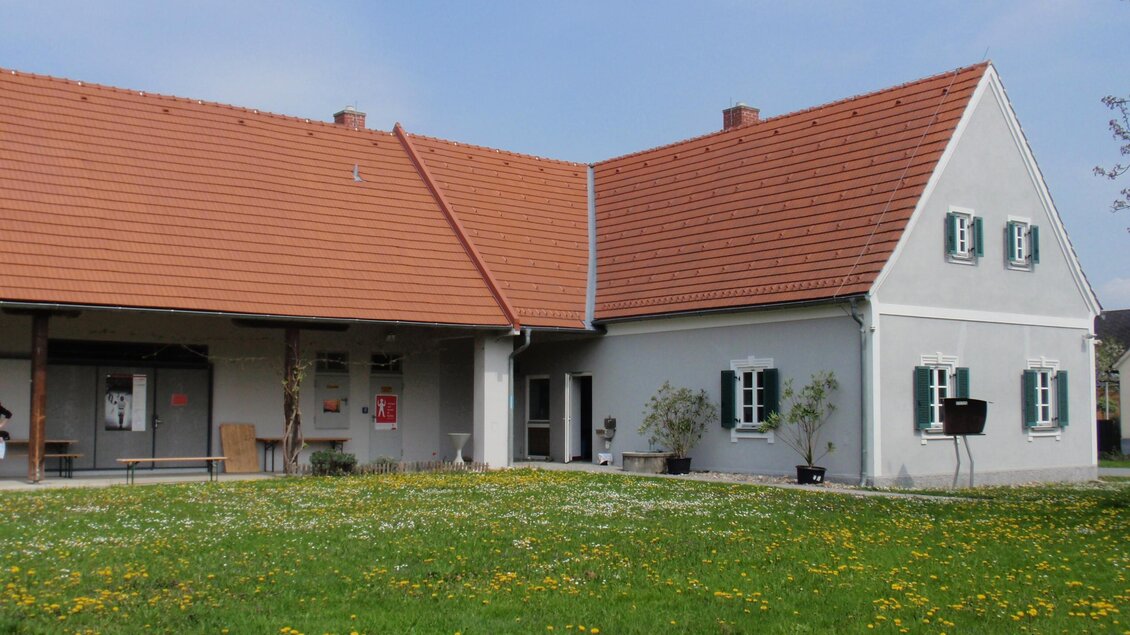 Ein modernes Haus mit einem rotem Dach und weißen Wänden. Der Garten ist grün und blühend, mit vielen Löwenzahnblumen. | © Pavelhaus