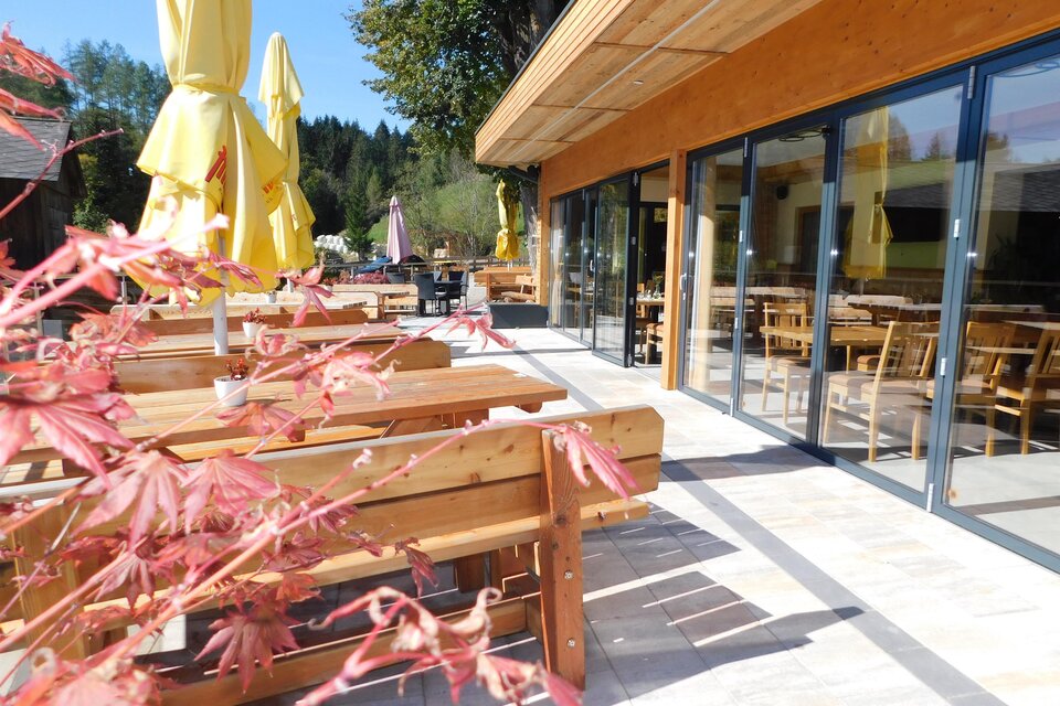 Ein einladender Außenbereich eines Restaurants mit Holzstühlen und Gelben Schirmen. Im Hintergrund sind große Fenster und ein schöner Ausblick auf die Natur.