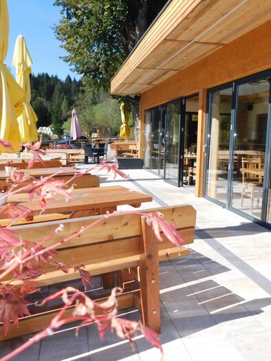 Ein einladender Außenbereich eines Restaurants mit Holzstühlen und Gelben Schirmen. Im Hintergrund sind große Fenster und ein schöner Ausblick auf die Natur.