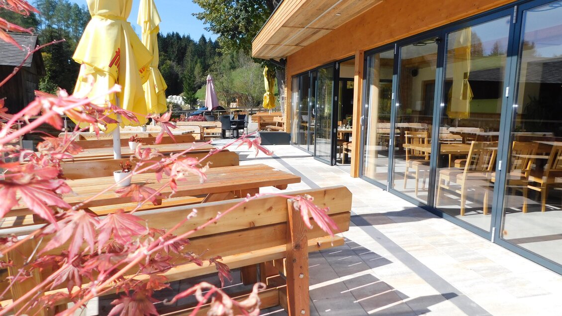 Ein einladender Außenbereich eines Restaurants mit Holzstühlen und Gelben Schirmen. Im Hintergrund sind große Fenster und ein schöner Ausblick auf die Natur.