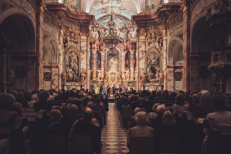 Kammermusikensemble spielt vor Publikum in einer barocken Kirche mit reich verziertem Altar und Deckenfresken. | © Reithofer Media