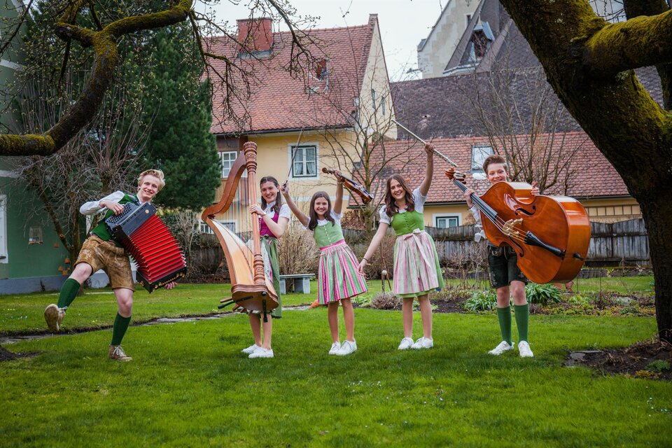Eine Gruppe von fünf jungen Musikern steht fröhlich in einem Garten. Sie spielen traditionelle Instrumente und tragen bunte Trachten. | © Zaumgspüt