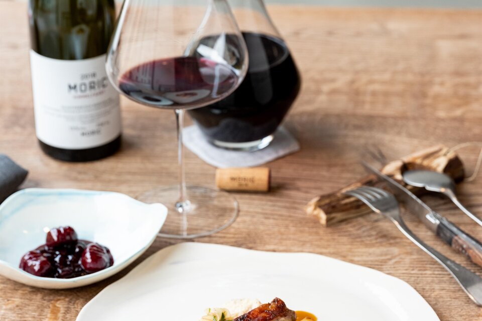 Ein elegantes Abendessen mit gebratenem Fleisch, Wurzelgemüse und einer Sauce auf einem weißen Teller. Dazu ein Glas Rotwein und eine Karaffe auf einem Holztisch. | © Michael Reidinger