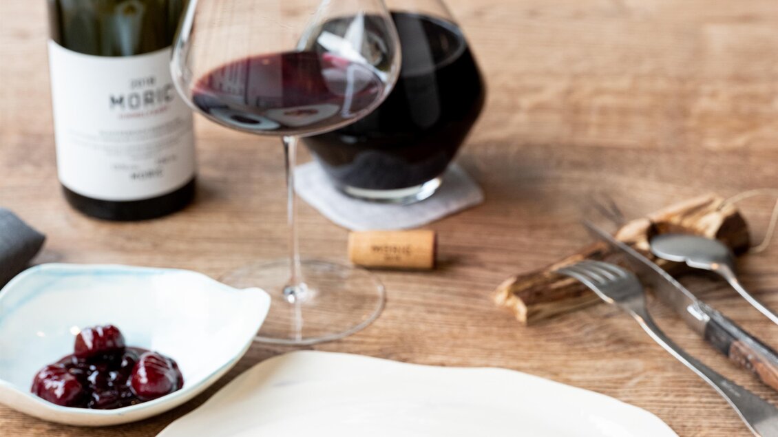 Ein elegantes Abendessen mit gebratenem Fleisch, Wurzelgemüse und einer Sauce auf einem weißen Teller. Dazu ein Glas Rotwein und eine Karaffe auf einem Holztisch. | © Michael Reidinger