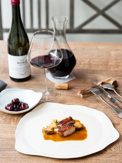 Ein elegantes Abendessen mit gebratenem Fleisch, Wurzelgemüse und einer Sauce auf einem weißen Teller. Dazu ein Glas Rotwein und eine Karaffe auf einem Holztisch. | © Michael Reidinger