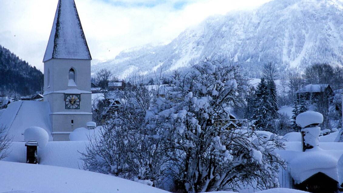 Ein verschneites Dorf mit einem Glockenturm und einem großen Baum. Die Berge im Hintergrund sind ebenfalls mit Schnee bedeckt. | © TVB Ausseerland Salzkammergut_Pirker