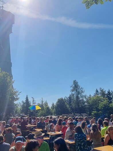 Ein belebter Ort mit vielen Menschen, die an Tischen sitzen. Im Hintergrund steht ein großer Turm und der Himmel ist klar und sonnig. | © Oststeiermark Tourismus