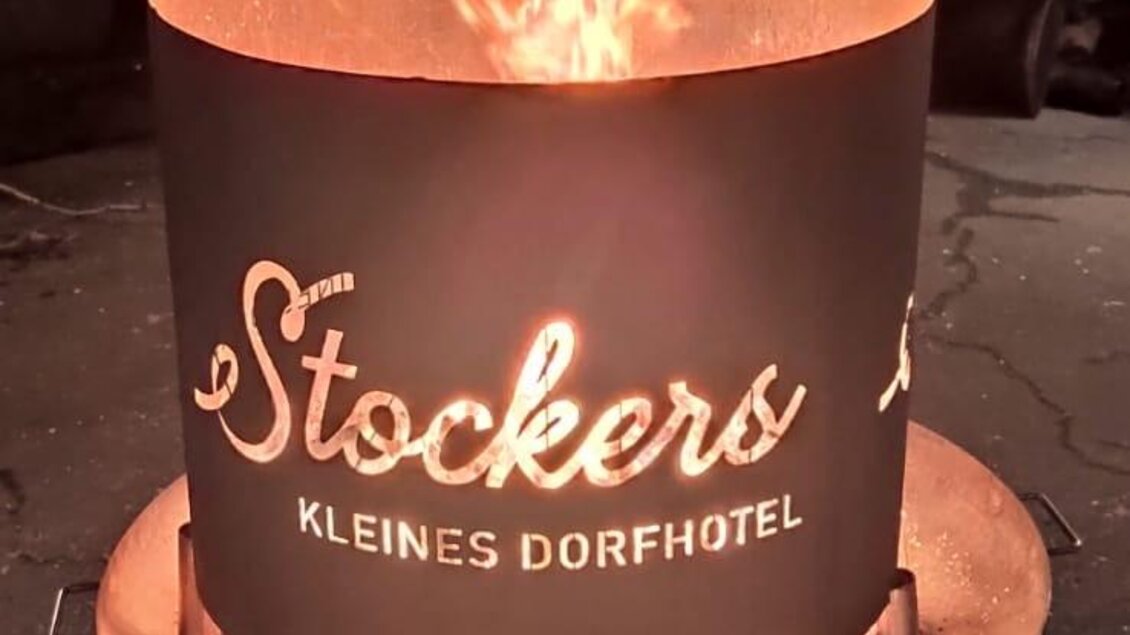 Ein Feuer in einer runden Feuerschale mit dem Schriftzug "Stockers - Kleines Dorfhotel". Funken fliegen und erzeugen eine gemütliche Atmosphäre. | © Peter Stocker