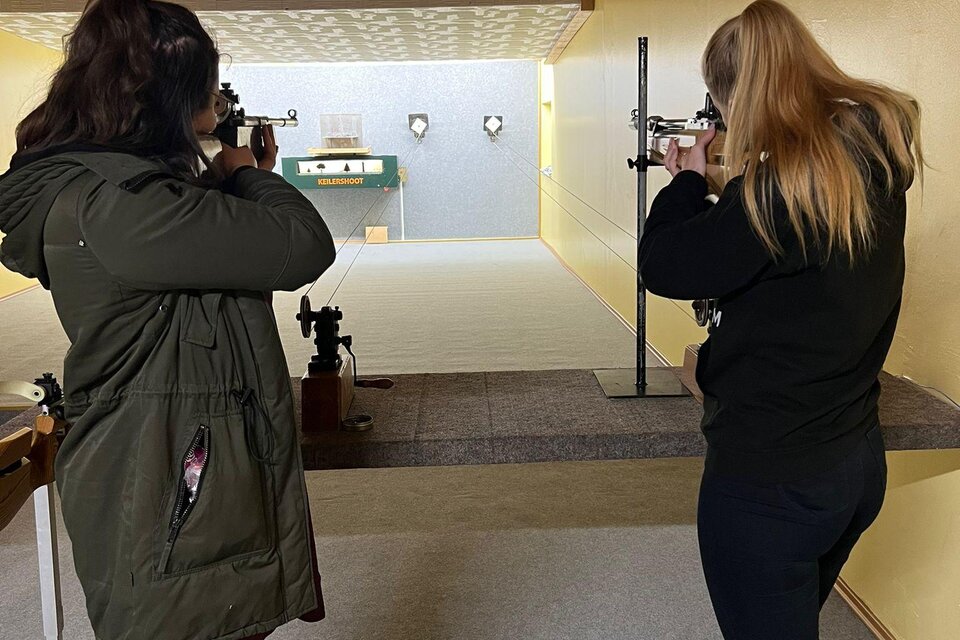 Zwei Frauen stehen in einer Schießhalle und zielen mit Gewehren auf ein Ziel. Der Raum hat eine helle Wandfarbe und eine Holzdecke. | © Tamara Neubauer