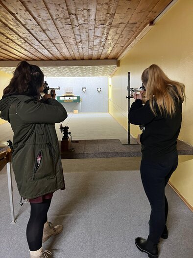 Zwei Frauen stehen in einer Schießhalle und zielen mit Gewehren auf ein Ziel. Der Raum hat eine helle Wandfarbe und eine Holzdecke. | © Tamara Neubauer