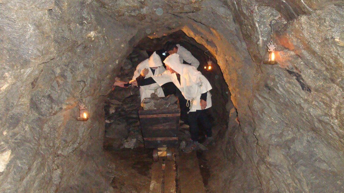 Ein schmaler Bergbau-Tunnel mit rauen Steinwänden. Im Hintergrund sind Lichtquellen und Holzbohlen sichtbar.