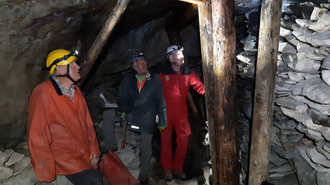 Drei Bergleute stehen in einem unterirdischen Stollen. Die Umgebung ist dunkel, mit Holzstützen und Steinmauern sichtbar.