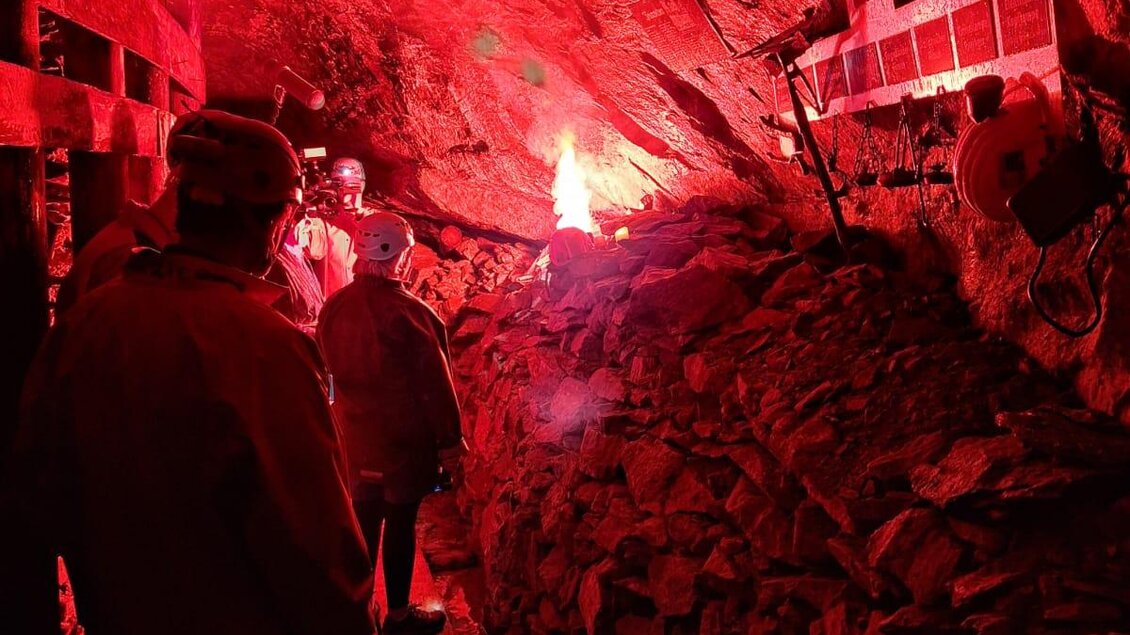 Ein unterirdischer Tunnel in rötlichem Licht mit Felsen und einer Holzplanke. Menschen stehen daneben und betrachten das Szenario.