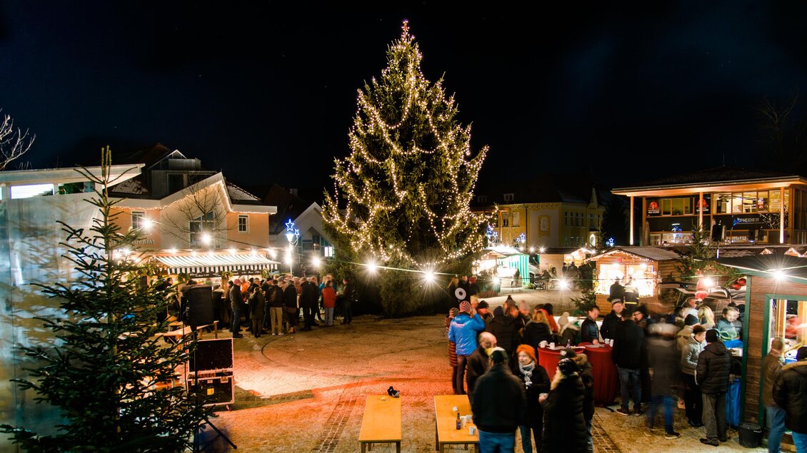Ein festlicher Weihnachtsmarkt bei Nacht mit einem großen, beleuchteten Weihnachtsbaum. Menschen stehen um Stände mit Essen und Getränken versammelt. | © TV Region Graz-Mias Photoart