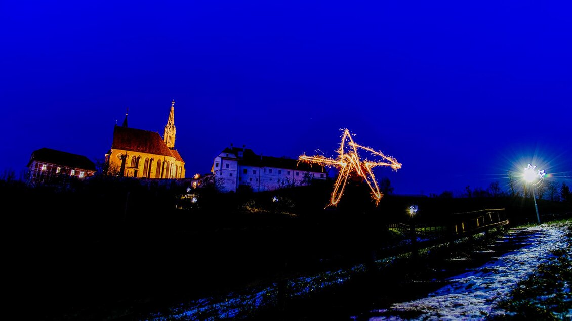 Eine festlich beleuchtete Kirche steht neben einem historischen Gebäude im Abendlicht. Im Vordergrund ist ein Weg mit Schnee und Laternen zu sehen. | © TV Region Graz-Mias Photoart