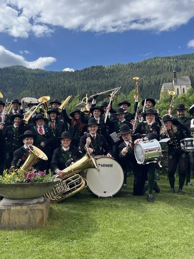 A large music group is outdoors, surrounded by green landscape and mountains. The musicians are wearing traditional clothing and holding their instruments high.