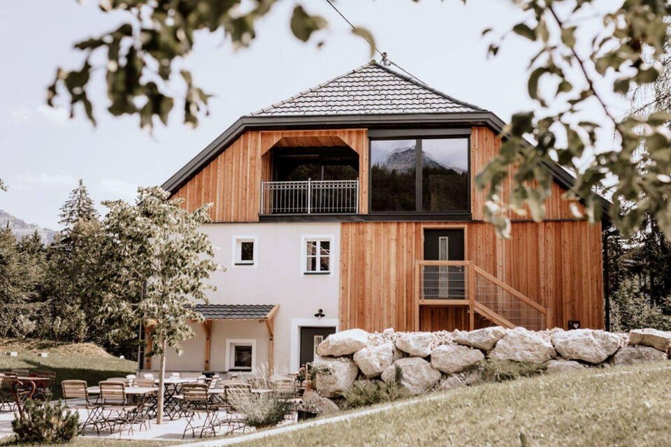 Die moderne Eingangsseite des Schutzhaus am hohen Camper in Bad Aussee mit Holzverkleidung und großen Fenstern in der naturnahen Umgebung des Ausseerlandes. Im Vordergrund sind Terrassenstühle und Tische zwischen kleinen Bäumen aufgestellt. | © Das Schutzhaus