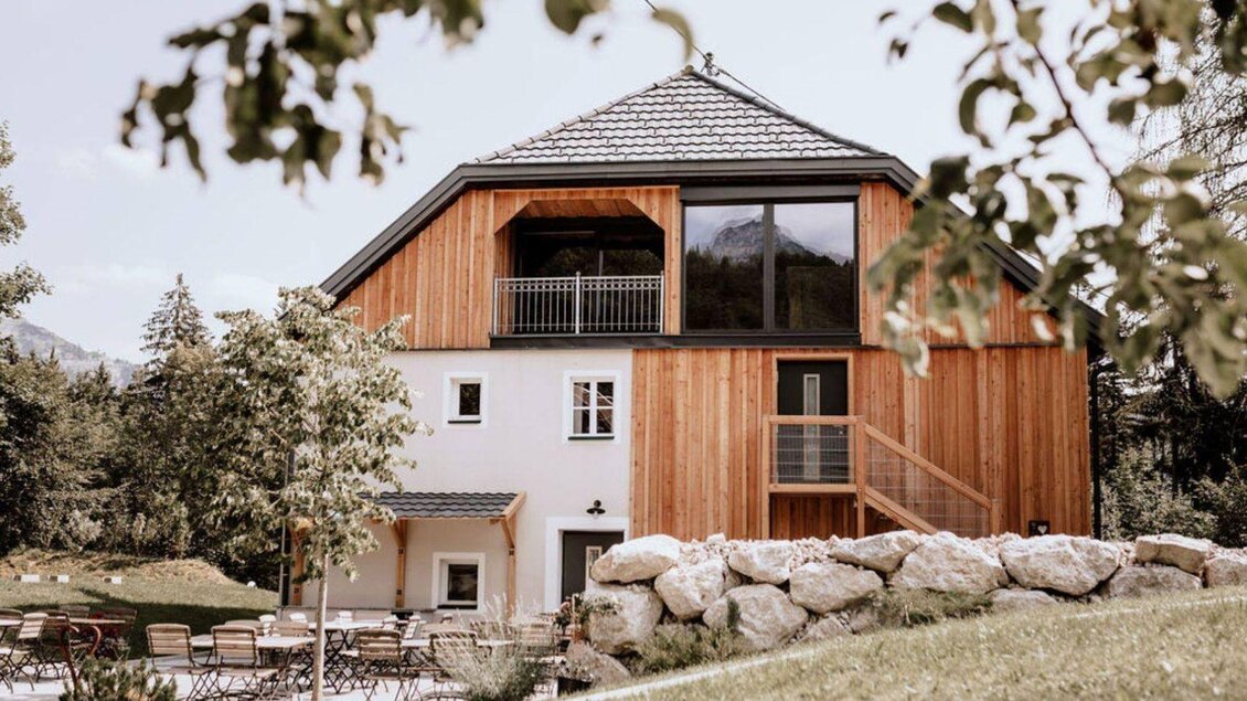 Die moderne Eingangsseite des Schutzhaus am hohen Camper in Bad Aussee mit Holzverkleidung und großen Fenstern in der naturnahen Umgebung des Ausseerlandes. Im Vordergrund sind Terrassenstühle und Tische zwischen kleinen Bäumen aufgestellt. | © Das Schutzhaus