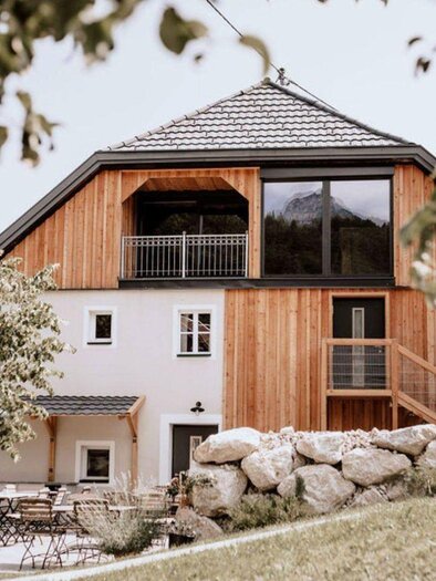 The modern entrance of the hotel "Schutzhaus am hohen Camper" in Bad Aussee with wooden cladding and large windows in the nature-friendly surroundings of the Ausseerland region. In the foreground, terrace chairs and tables are set up among small trees. | © Das Schutzhaus