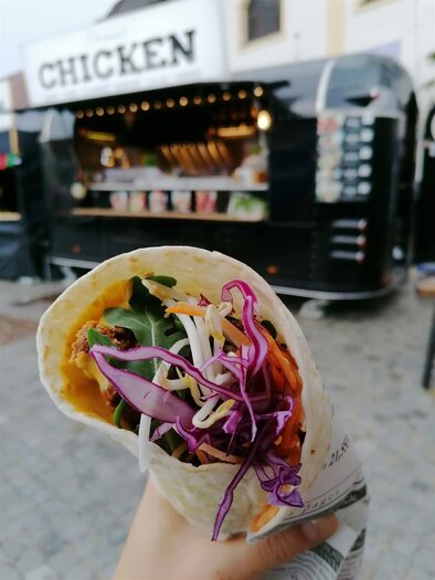 Egy kézben tartott wrap friss zöldségekkel és mártással. A háttérben egy étkezde látható, amelyen a "Chicken" felirat olvasható. | © Tourismusverband Oststeiermark