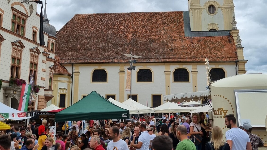 Ein belebter Markt mit vielen Menschen und Ständen. Im Hintergrund ist eine Kirche mit einer markanten Uhr zu sehen. | © Grill Thomas