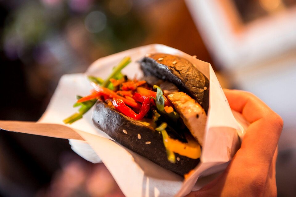 Ein handgehaltener Burger mit schwarzem Brötchen, gefüllt mit Gemüse und einer Fleischscheibe. Die bunten Zutaten sorgen für einen appetitlichen Anblick. | © Street Food Market Austria