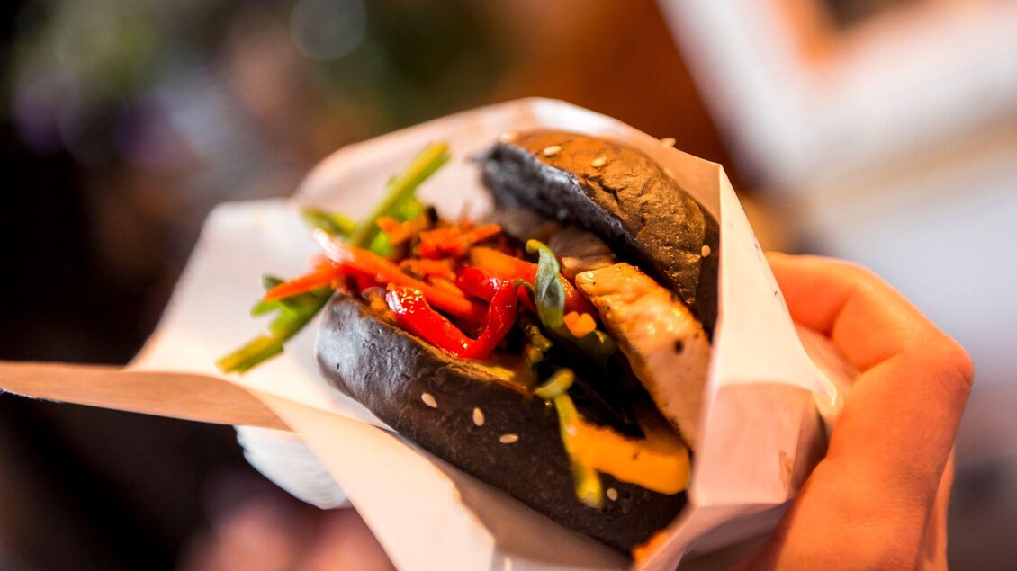 Ein handgehaltener Burger mit schwarzem Brötchen, gefüllt mit Gemüse und einer Fleischscheibe. Die bunten Zutaten sorgen für einen appetitlichen Anblick. | © Street Food Market Austria